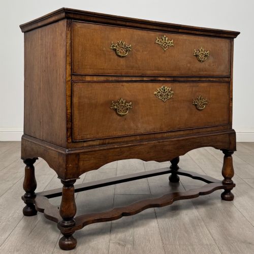fig 7. An antique 19th-century William and Mary style walnut chest of drawers featuring two deep drawers with ornate brass drop handles, raised on turned legs connected by a wavy stretcher base.