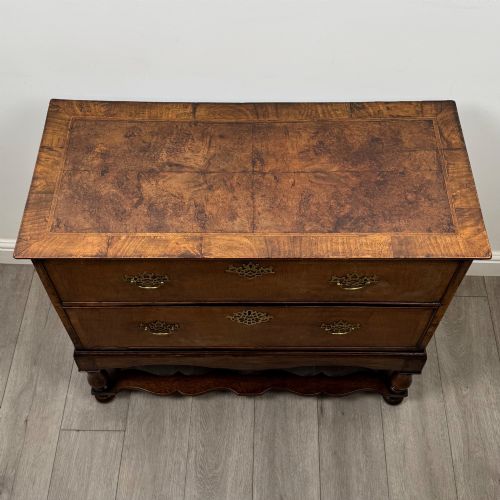 fig 8. An antique 19th-century William and Mary style walnut chest of drawers featuring two deep drawers with ornate brass drop handles, raised on turned legs connected by a wavy stretcher base.