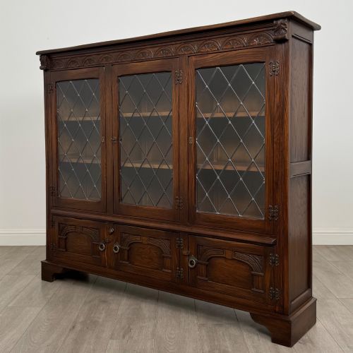 fig 2. A large dark brown carved oak display cabinet featuring three leaded glass doors over three carved cupboard doors with metal ring pulls.