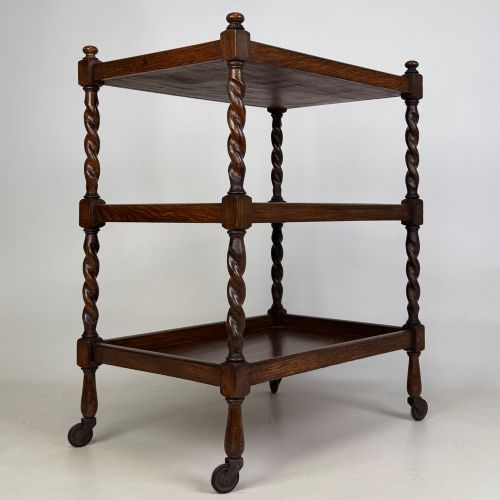 fig 3. A three-tier dark oak tea trolley featuring spiralled barley twist corner supports, turned top finials, and castor wheels.