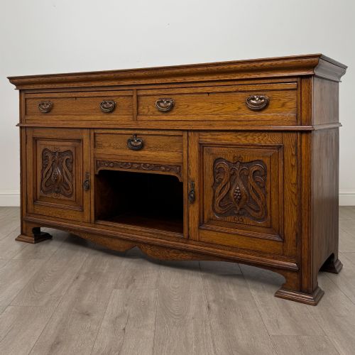 fig 2. An early 20th-century oak Arts and Crafts sideboard featuring hand-carved foliate panels, copper drop handles, multiple drawers, and a central open recess.