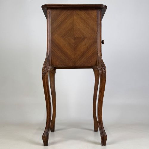 fig 7. An antique French walnut bedside cabinet featuring high slender cabriole legs, a single drawer, and a carved wooden door with a marquetry panel.