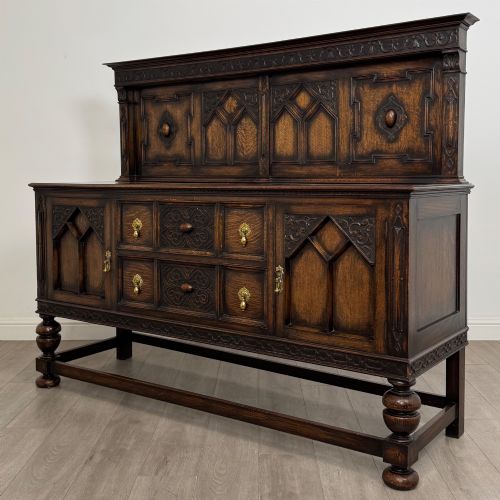 fig 2. A dark oak Jacobean style sideboard featuring a raised upper superstructure, geometric carved panelling, drawers with brass drop handles, and turned bulbous legs connected by a stretcher base.