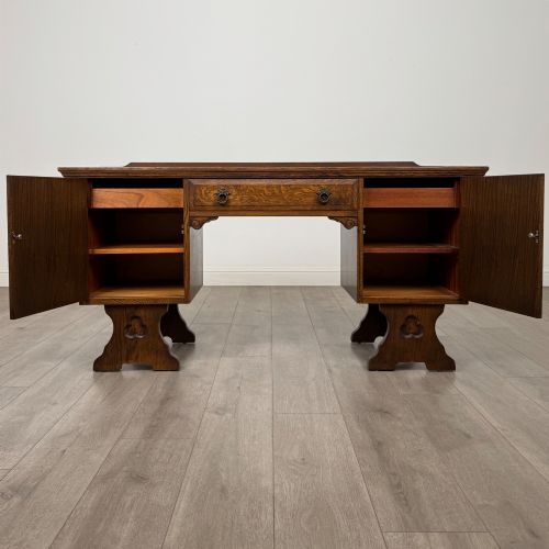 fig 1. A 20th-century carved oak pedestal desk featuring linenfold cupboard doors, a central top drawer, and trefoil pierced end supports.