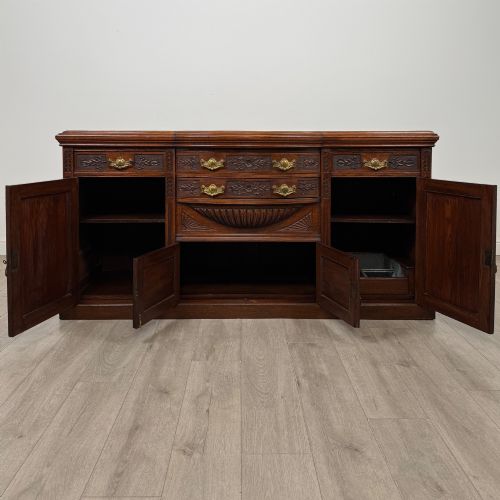 fig 1. A wide 19th-century Victorian dark oak sideboard featuring relief-carved floral panels, central drawers with brass handles, and lower cupboard doors.