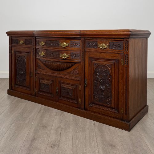 fig 2. A wide 19th-century Victorian dark oak sideboard featuring relief-carved floral panels, central drawers with brass handles, and lower cupboard doors.