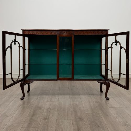 fig 1. An early twentieth-century mahogany display cabinet featuring three glazed doors with curved astragal glazing bars, a carved top frieze, green fabric interior, and cabriole legs.