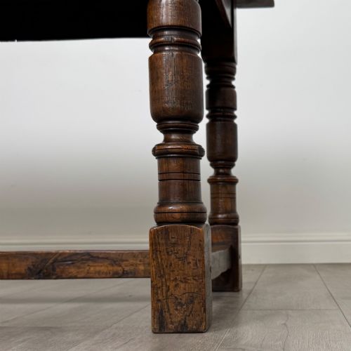 fig 5. A dark carved oak refectory dining table featuring a carved frieze apron, turned baluster legs, and an H-stretcher base on light timber flooring.