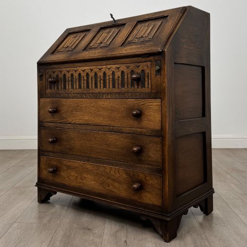 fig 2. A dark oak writing bureau with a slanted fall-front upper section featuring three carved linenfold panels above four drawers with turned wooden knob handles.