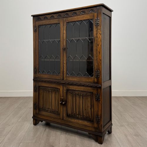 fig 2. A dark oak display cabinet featuring two upper leaded glass doors, lower cupboard doors carved with linenfold panels, and decorative metal hinges.