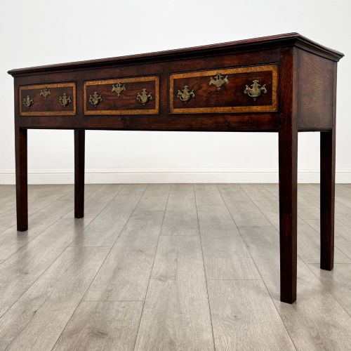 fig 1. An antique dark oak dresser base featuring three crossbanded drawers with brass drop handles and keyhole escutcheons, supported by four straight square legs.