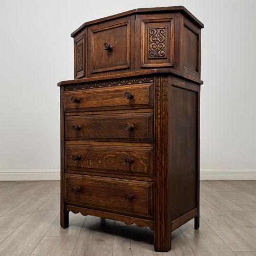 fig 2. An Edwardian dark oak cupboard featuring an upper canted cabinet with carved decorative panels above four graduated drawers with turned wooden knobs.