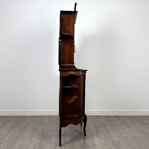 fig 7. A late 19th-century mahogany display cabinet featuring floral marquetry inlay, glazed central doors, oval side mirrors, and a decorative asymmetric crest pediment.