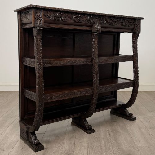 fig 7. A 19th-century dark oak open bookcase featuring hand-carved frieze foliage, a central mask motif, and three tiers of open shelving.