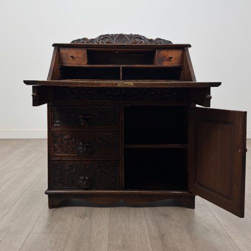 fig 1. A dark oak Victorian slant-front bureau featuring carved Green Man drawer pulls, foliage relief carvings, and a single panelled cupboard door.