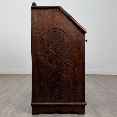 fig 7. A dark oak Victorian slant-front bureau featuring carved Green Man drawer pulls, foliage relief carvings, and a single panelled cupboard door.
