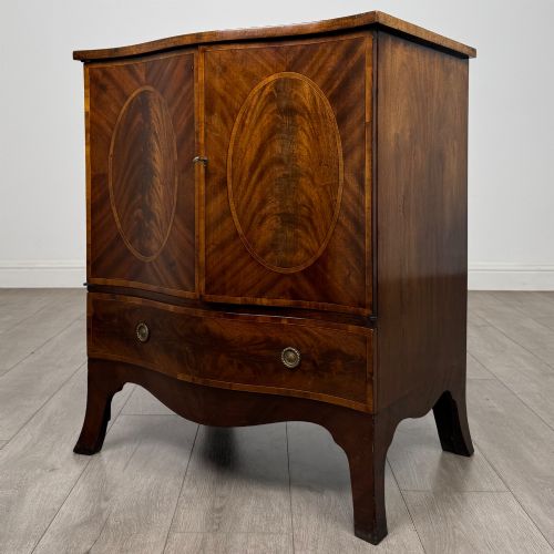 fig 2. A 20th-century mahogany serpentine cupboard featuring flame mahogany oval door panels, chevron veneering, a lower drawer with brass ring handles, and splayed bracket feet.