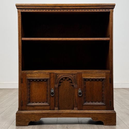 fig 1. A small medium-brown oak open bookcase featuring two upper shelves and a lower two-door cupboard with carved arched motifs and metal ring handles.