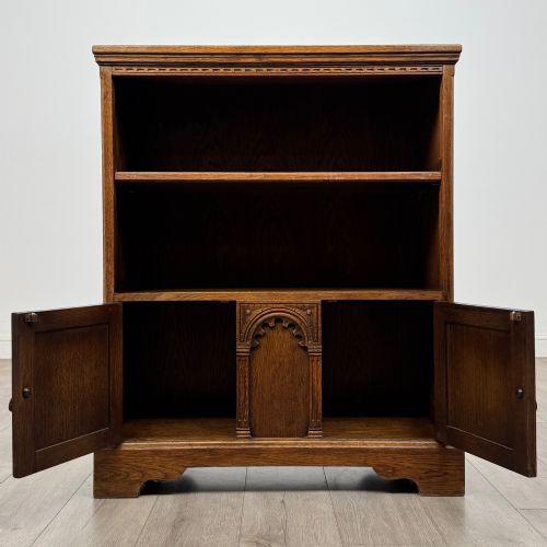fig 2. A small medium-brown oak open bookcase featuring two upper shelves and a lower two-door cupboard with carved arched motifs and metal ring handles.