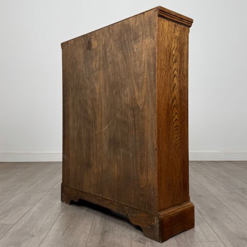 fig 7. A small medium-brown oak open bookcase featuring two upper shelves and a lower two-door cupboard with carved arched motifs and metal ring handles.