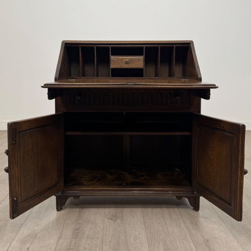 fig 1. A dark oak slant-front writing bureau featuring carved linenfold upper panels, a single carved drawer with wooden knobs, and a double-door cupboard base decorated with diamond lozenge carvings.