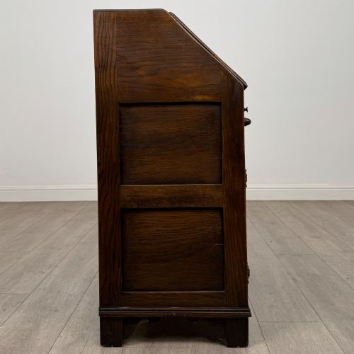 fig 7. A dark oak slant-front writing bureau featuring carved linenfold upper panels, a single carved drawer with wooden knobs, and a double-door cupboard base decorated with diamond lozenge carvings.