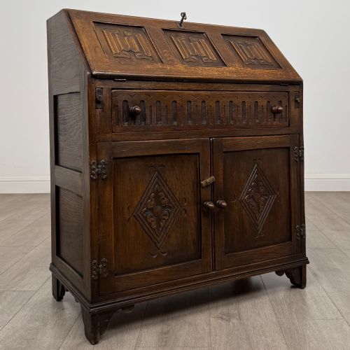 fig 8. A dark oak slant-front writing bureau featuring carved linenfold upper panels, a single carved drawer with wooden knobs, and a double-door cupboard base decorated with diamond lozenge carvings.