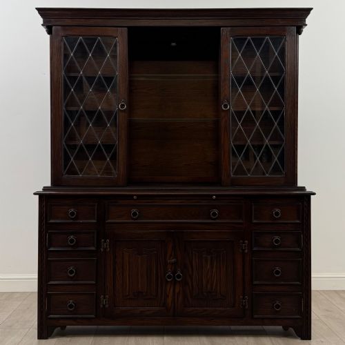 fig 1. A dark oak display cabinet with a leaded glass central upper cupboard, flanked by open shelves, set atop a lower unit with multiple drawers and carved cupboard doors.