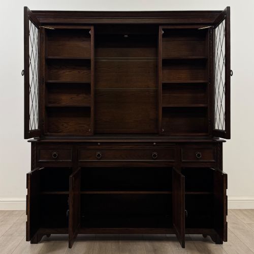 fig 2. A dark oak display cabinet with a leaded glass central upper cupboard, flanked by open shelves, set atop a lower unit with multiple drawers and carved cupboard doors.