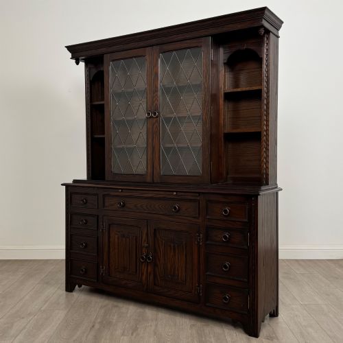 fig 3. A dark oak display cabinet with a leaded glass central upper cupboard, flanked by open shelves, set atop a lower unit with multiple drawers and carved cupboard doors.