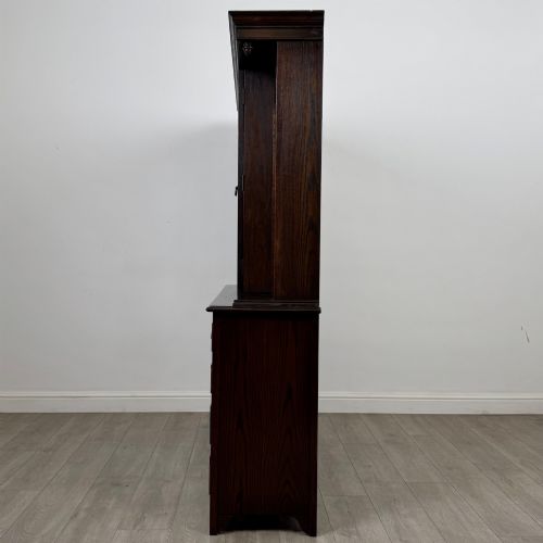 fig 4. A dark oak display cabinet with a leaded glass central upper cupboard, flanked by open shelves, set atop a lower unit with multiple drawers and carved cupboard doors.