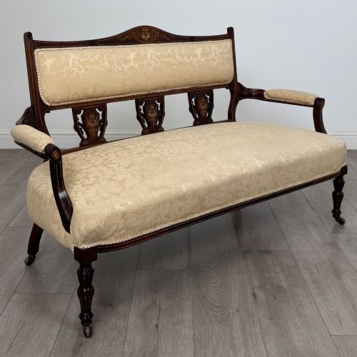 fig 7. An antique Victorian rosewood settee featuring central backrest marquetry inlay, open armrests, turned front legs on castors, and cream damask upholstery.