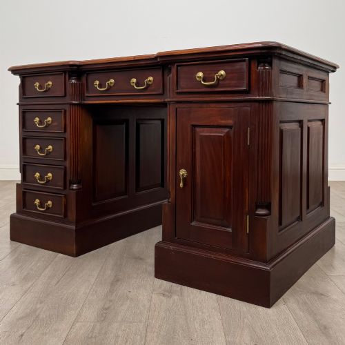 fig 2. A dark mahogany pedestal desk featuring a central kneehole drawer flanked by five drawers on the left and a panelled cupboard door on the right, all fitted with brass drop handles.