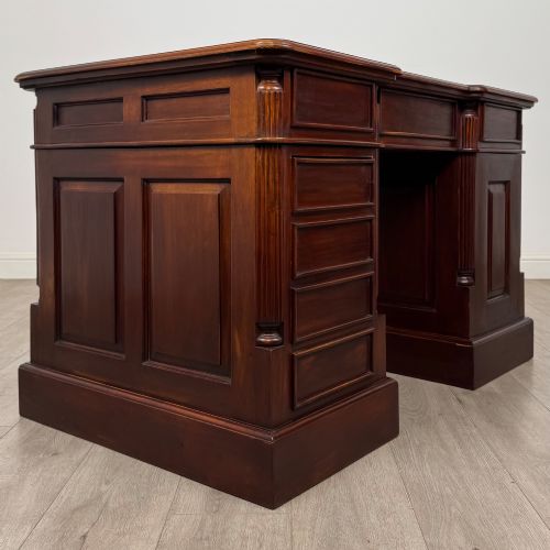 fig 4. A dark mahogany pedestal desk featuring a central kneehole drawer flanked by five drawers on the left and a panelled cupboard door on the right, all fitted with brass drop handles.