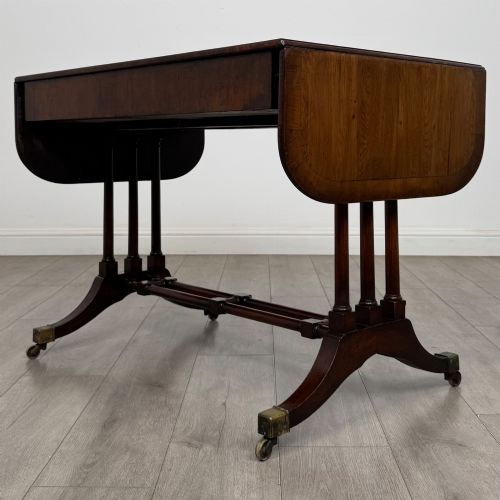 fig 6. A 19th-century Regency style yew wood side table featuring drop-leaf ends, twin drawers with brass escutcheons and round knobs, turned twin-stem supports, a stretcher, and brass castors.