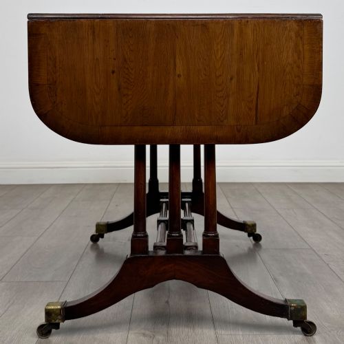 fig 7. A 19th-century Regency style yew wood side table featuring drop-leaf ends, twin drawers with brass escutcheons and round knobs, turned twin-stem supports, a stretcher, and brass castors.