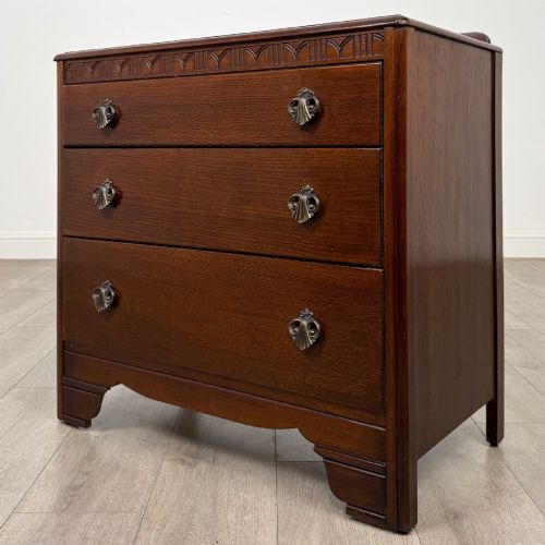 fig 1. A dark brown oak chest of drawers featuring three wide drawers with ornamental metal drop handles and a carved geometric frieze along the top edge.
