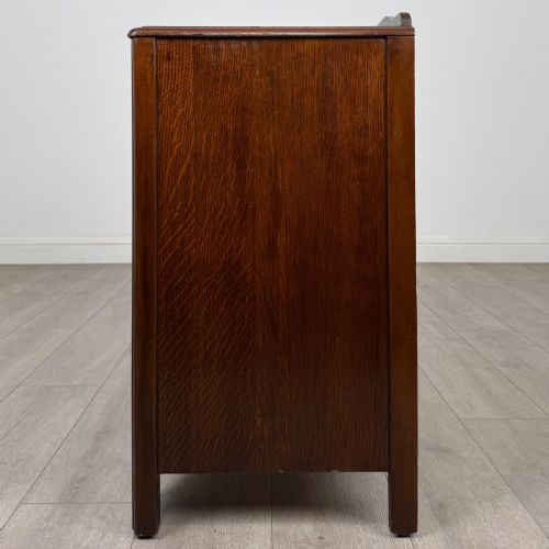 fig 2. A dark brown oak chest of drawers featuring three wide drawers with ornamental metal drop handles and a carved geometric frieze along the top edge.