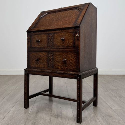 fig 2. A small Edwardian dark oak writing bureau on a tall stand with two drawers featuring carved central panels and turned knobs.
