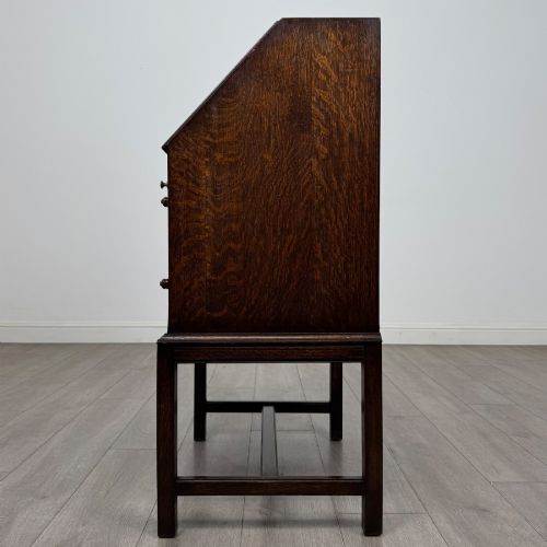fig 3. A small Edwardian dark oak writing bureau on a tall stand with two drawers featuring carved central panels and turned knobs.