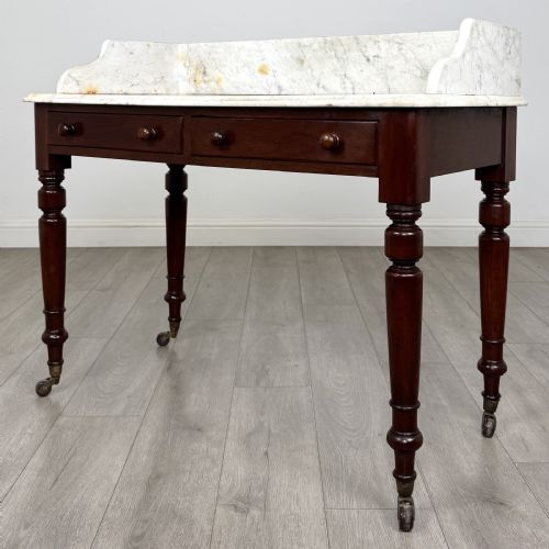 fig 1. A Victorian mahogany washstand featuring a white marble top with a raised splashback, two drawers with turned knobs, turned legs, and brass castors.