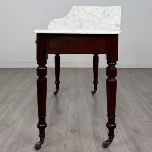fig 2. A Victorian mahogany washstand featuring a white marble top with a raised splashback, two drawers with turned knobs, turned legs, and brass castors.