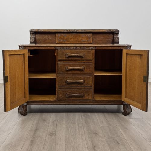fig 1. An antique golden oak Art Deco sideboard featuring a carved gallery back, four central drawers, flanked cupboard doors with carved floral panels, and sculpted wooden handles.