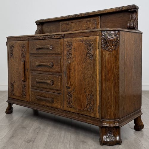 fig 2. An antique golden oak Art Deco sideboard featuring a carved gallery back, four central drawers, flanked cupboard doors with carved floral panels, and sculpted wooden handles.