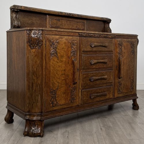 fig 8. An antique golden oak Art Deco sideboard featuring a carved gallery back, four central drawers, flanked cupboard doors with carved floral panels, and sculpted wooden handles.