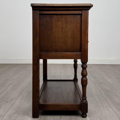 fig 6. A dark oak side table featuring five drawers with brass handles, turned front legs, an arched kneehole, and a lower tier shelf.