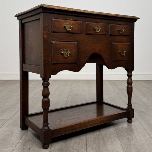 fig 7. A dark oak side table featuring five drawers with brass handles, turned front legs, an arched kneehole, and a lower tier shelf.