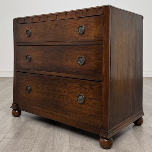 fig 1. A dark oak chest of three drawers featuring carved decorative detailing along the top edge, circular ring handles, and turned bun feet.