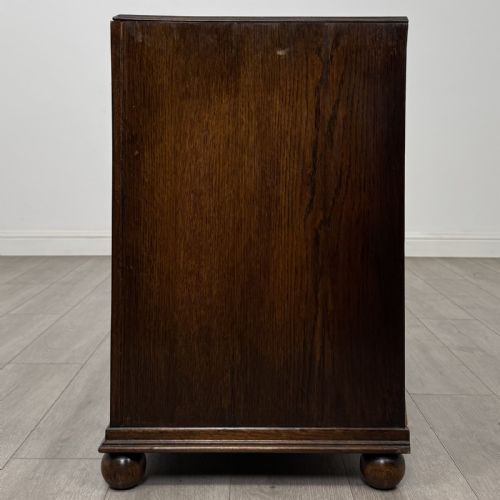 fig 2. A dark oak chest of three drawers featuring carved decorative detailing along the top edge, circular ring handles, and turned bun feet.