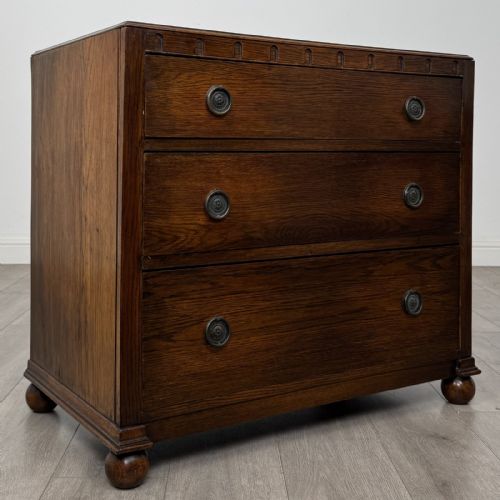 fig 7. A dark oak chest of three drawers featuring carved decorative detailing along the top edge, circular ring handles, and turned bun feet.